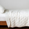 Harlow Coverlet | Winter White | Quilted cotton velvet coverlet, folded back to showcase linen back, draped over white sheeting - side view.