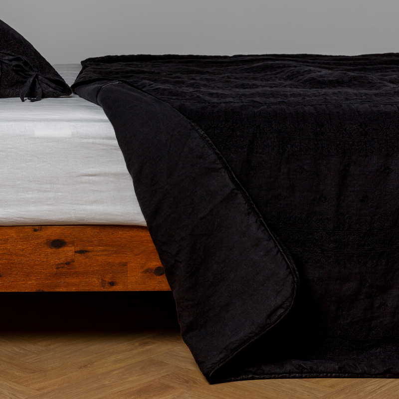 Ines Bedspread | Corvino | embroidered bedspread with corner pulled back  showing the plain linen back. Shown with a white fitted sheet and matching sham, the bedframe is slightly visible  and the bed is against a white wall and on a medium wood floor.