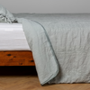 Ines Bedspread | Eucalyptus | embroidered bedspread with corner pulled back  showing the plain linen back. Shown with a white fitted sheet and matching sham, the bedframe is slightly visible  and the bed is against a white wall and on a medium wood floor.