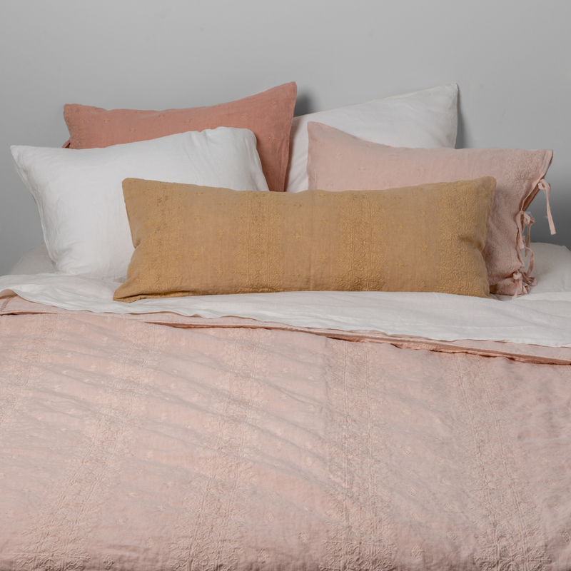 Ines Bedspread | Pearl | Embroidered bedspread shown in pearl with white sheets and  sleping pillows, and warm toned shams and throw pillow. Shot from the end of the bed and the sheet is pulled back over the top of the  bedspread.