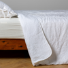 Ines Bedspread | White | embroidered bedspread with corner pulled back  showing the plain linen back. Shown with a white fitted sheet and matching sham, the bedframe is slightly visible  and the bed is against a white wall and on a medium wood floor.