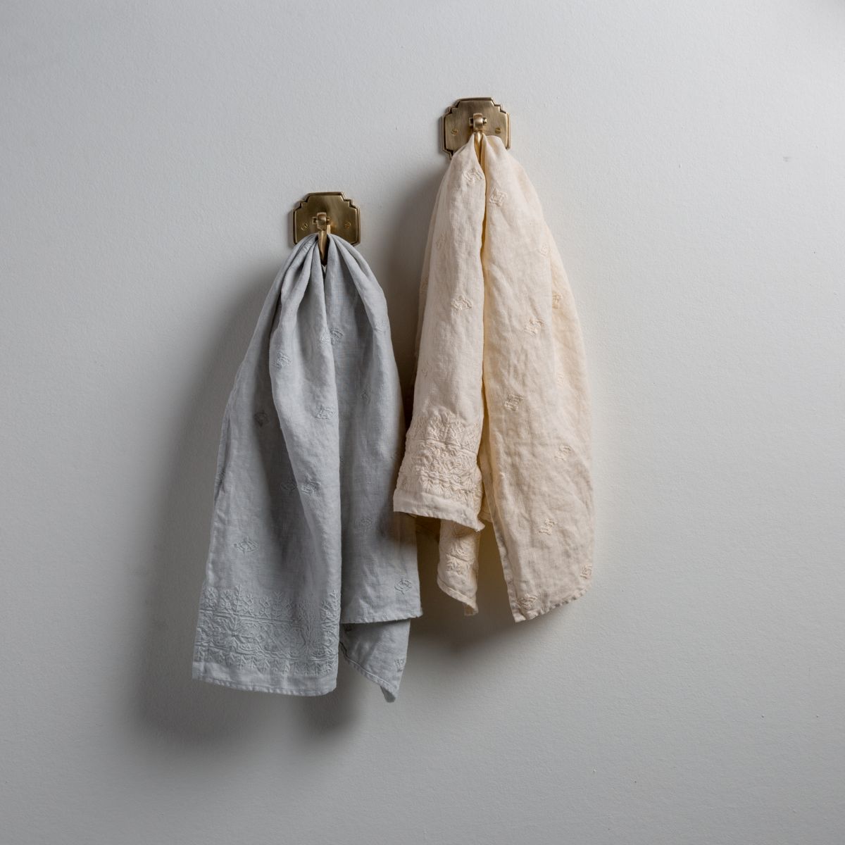 Two Ines guest towels, draped over decorative hooks against a white wall - cloud and parchment.