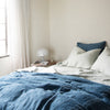Midnight Ines duvet cover layered with soft green linen sheets, partially unmade with light toned walls and curtains, all bathed in natural light - cropped three-quarter angle.