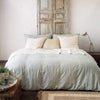 Ines duvet cover in eucalyptus with cream-toned sheets and pillows, in a room with distressed wood, light walls, and neutral accents - end of bed view.