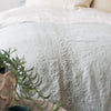 Ines Duvet Cover | Eucalyptus duvet cover on a bed with cream-toned sheeting, and greenery visible in the foreground - close-up end of bed view.