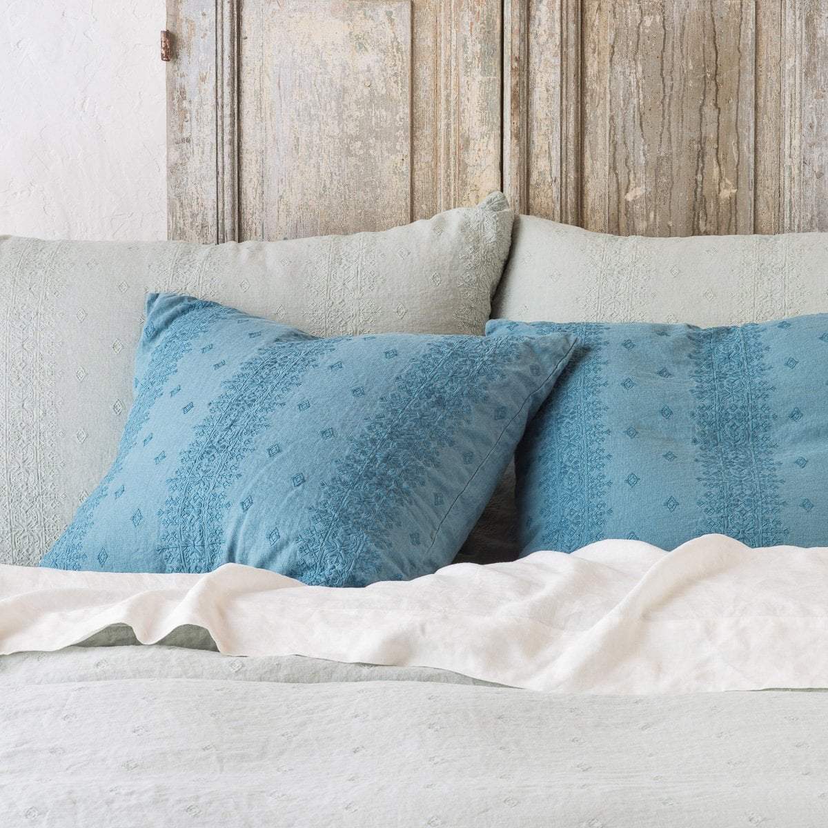 Eucalyptus and cenote Ines shams leaning upright against a distressed wood headboard on light toned bedding - cropped head-on view.