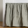 Linen Crib Skirt | Eucalyptus | crib skirt shown on a white crib with no mattress against a white wall and medium wood flooring.