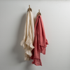 Two Linen Whisper guest towels, draped over decorative hooks against a white wall - parchment and poppy.