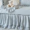 Linen crib sheet in cloud with matching crib skirt, baby blanket, and throw pillow on a white iron crib decorated with stuffed bear - side view.