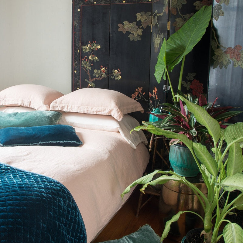 Linen duvet cover in pearl on a neatly made bed. Silk velvet accents in blues and greens are complemented by on-tone ceramics, dark floral wall panel, and tall greenery - cropped three-quarter angle.