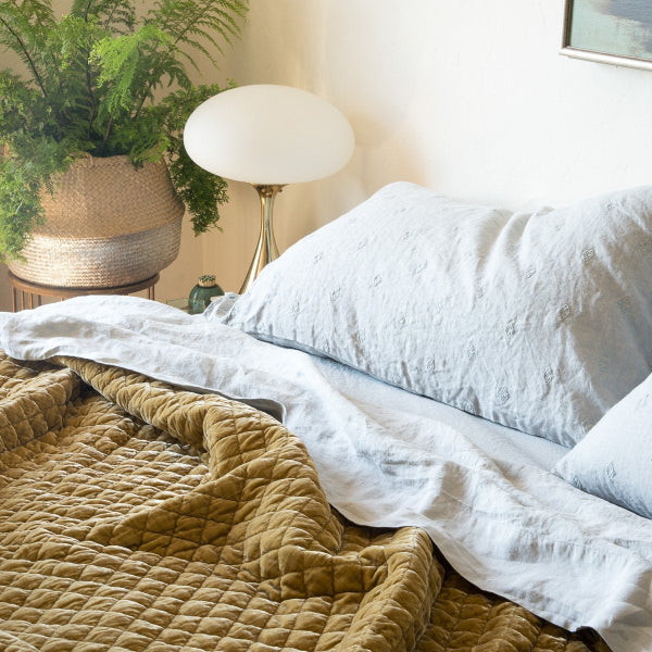 [allvariants]: Cloud linen sheeting with embroidered linen shams and rich gold toned silk velvet quilted coverlet, styled with a green fern in a woven basket and gold metal accents - cropped three-quarter angle.