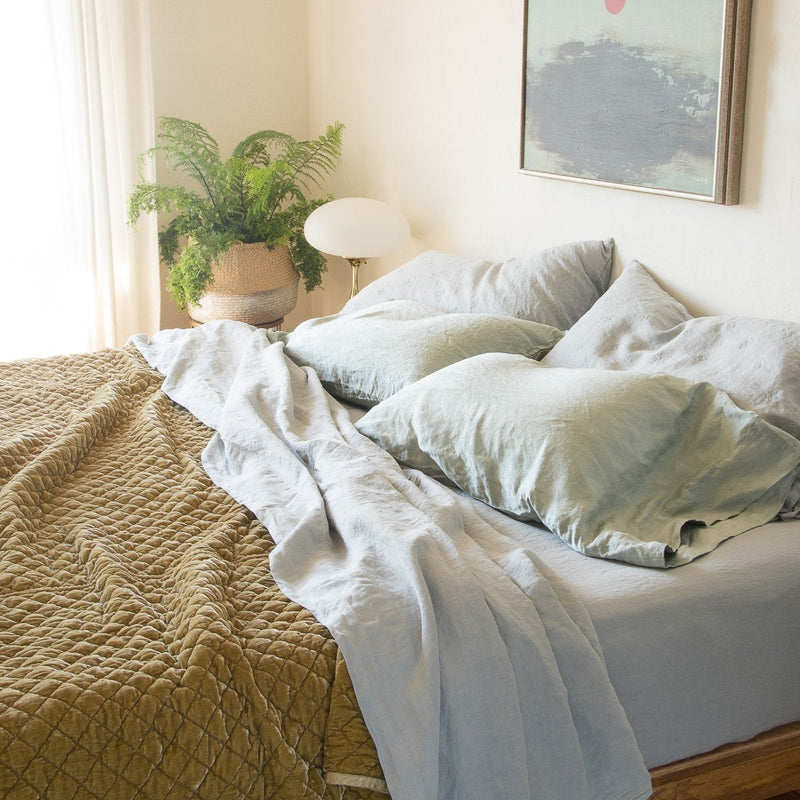 Linen Fitted Sheet | Linen sheeting in cloud with on-tone silk charmeuse sleeping pillows and rich gold toned silk velvet quilted coverlet, styled simply in natural light with a green fern and soft green art - cropped three-quarter angle.