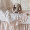 Linen Whisper Baby Blanket | Linen whisper baby comforter in pearl, draped over matching sheet and crib skirt, shown with a stuffed bunny and twinkly lights - side view.
