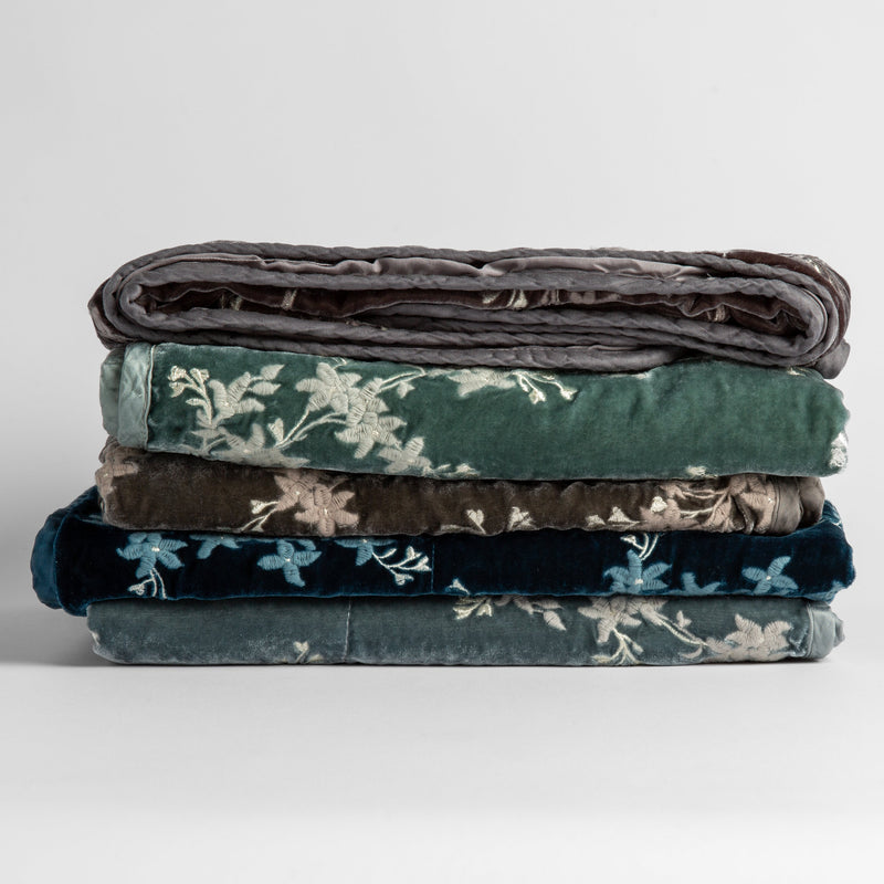 Lynette Baby Blanket | a stack of grey and green toned embroidered silk velvet baby blankets against a white background.