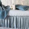 Madera Luxe crib sheet shown on a white iron crib with silk velvet throw blanket and pillow, all in cloud - side view.