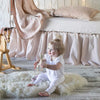 Madera Luxe crib sheet in pearl with on-tone baby blanket, pillow, and crib skirt on a white iron crib. Side view with baby sitting and playing in the foreground.