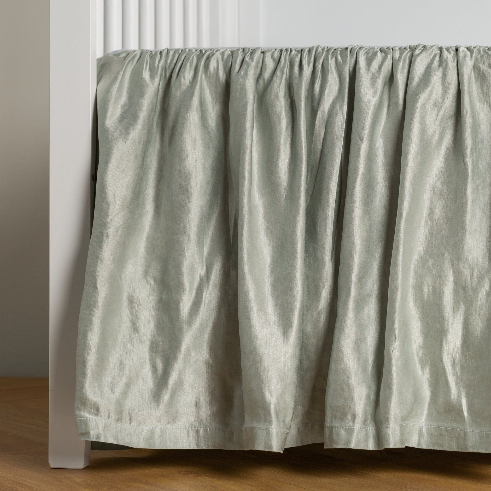 Paloma Crib Skirt | Cloud | charmeuse crib skirt shown on a white crib against a white wall and medium wood floor.