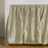 Paloma Crib Skirt | Parchment | charmeuse crib skirt shown on a white crib against a white wall and medium wood floor.