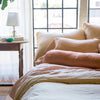Paloma Throw Pillow | Paloma bolster and sleeping pillows on linen bedding, in soft pink and gold tones against big windows - cropped end of bed view.