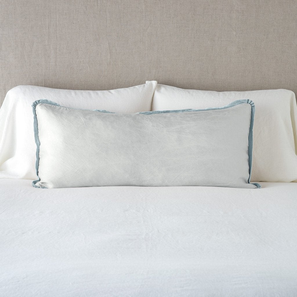 Paloma Throw Pillow | Cloud | 16x36 charmeuse pllow wth silk velvet trim against white sleeping pillows and sheets — straight on with neutral background.