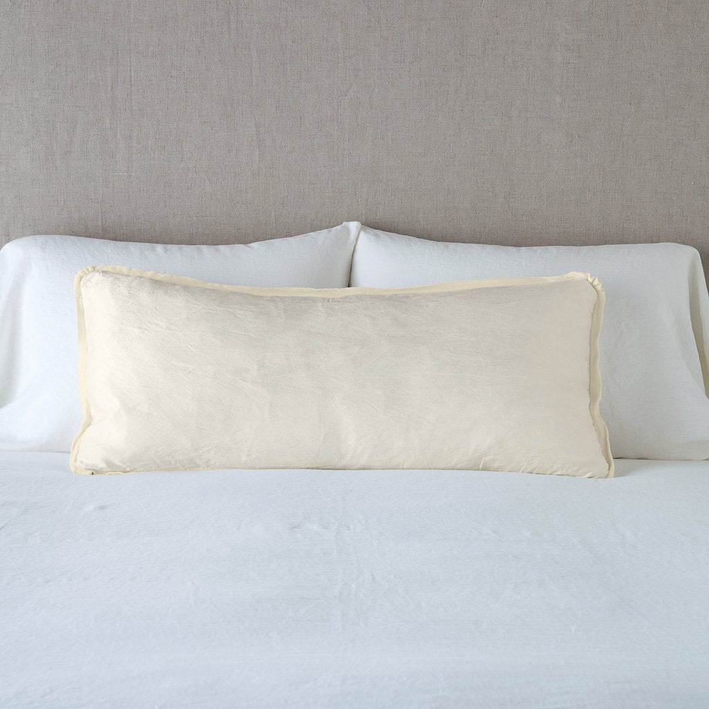 Paloma Throw Pillow | Parchment | 16x36 charmeuse pllow wth silk velvet trim against white sleeping pillows and sheets — straight on with neutral background.