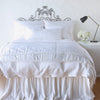 Paloma Blanket | White | throw blanket folded at the end of a monochromatic, charmeuse bed - end of bed view.