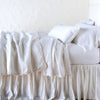 Paloma Blanket | White | throw blanket rumpled on a charmeuse monochromatic bed - side view.