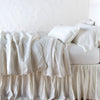 Paloma Blanket | Winter White | throw blanket rumpled on a charmeuse monochromatic bed - side view.