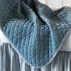 Close up of silk velvet quilted baby blanket in cloud, draped over a monochromatic crib, showcasing the satin edge trim.