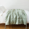 Taline Blanket | Eucalyptus | Taline throw blanket draped over a white bed - eucalyptus, side view.