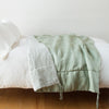Taline Blanket | Eucalyptus | blanket draped over a white bed, folded back to reveal midweight linen back - side view.