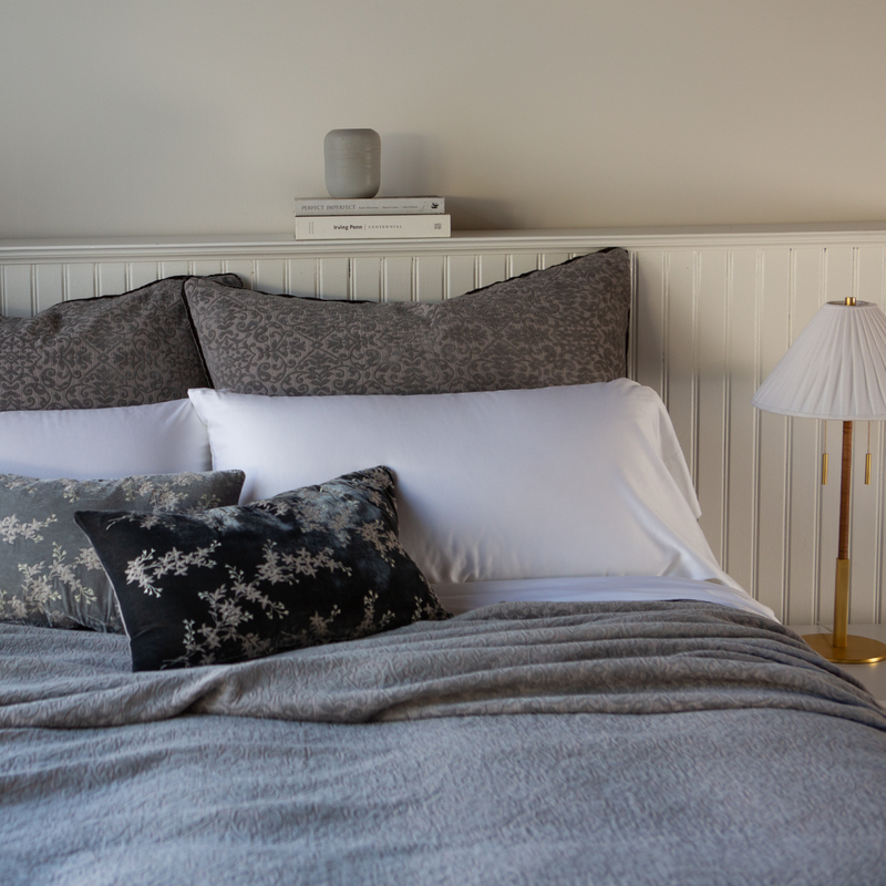 Vienna Twin Coverlet | Moonlight | cotton chenile coverlet with matching shams shown from the foot of the bed with white sleeping pillows and grey decorative pillows. A lamp is on the side table and a short stack of books is on the beadboard shelf.